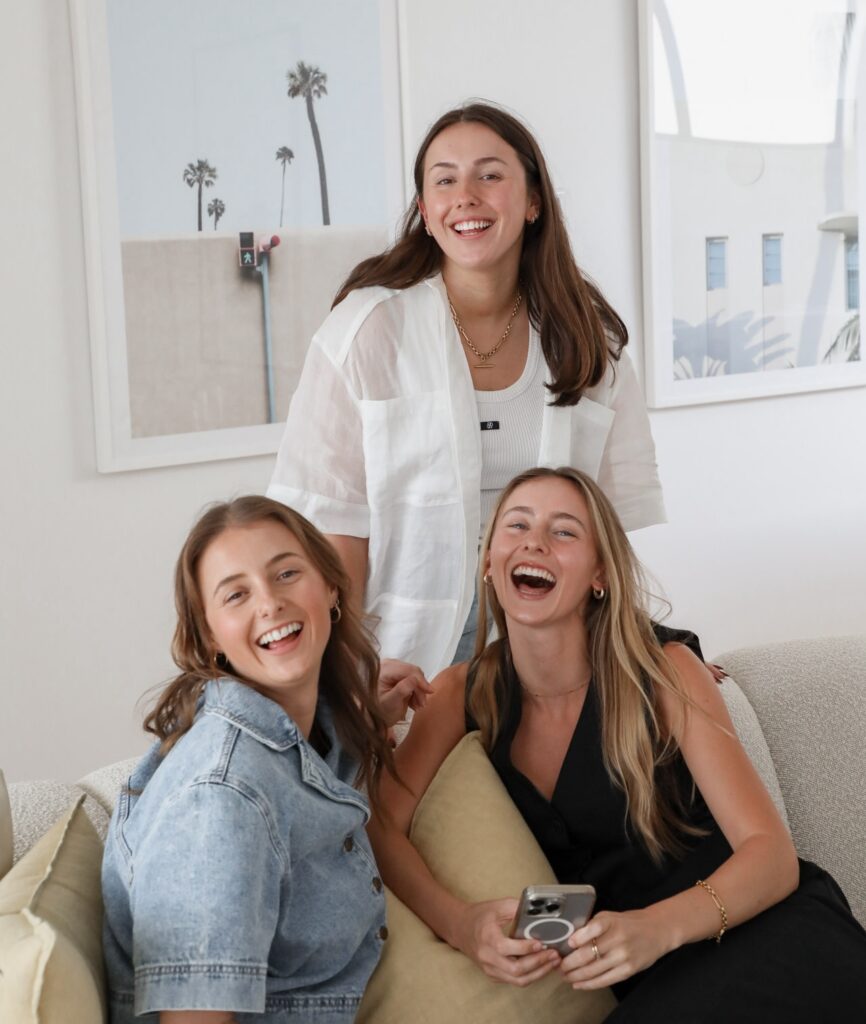 three girls sitting on a beige couch, laughing and smiling at the camera. One of the girls is holding an iPhone and there are coastal prints on the wall in the background